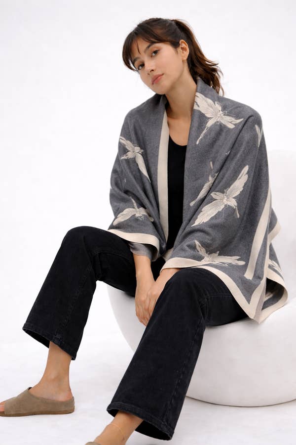 A woman sits on a white round stool wearing a gray open-front cardigan with a white dragonfly pattern, black cropped pants, and taupe slip-on shoes against a plain white studio background.