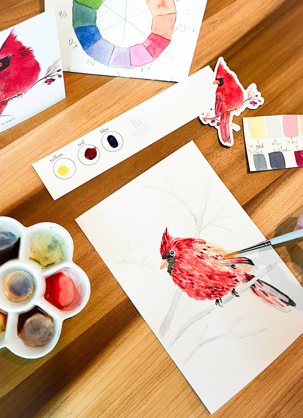 A watercolor painting of a red cardinal bird on white paper, shown on a wooden table with a paint palette, watercolor wheel, color swatches, and other painting supplies.