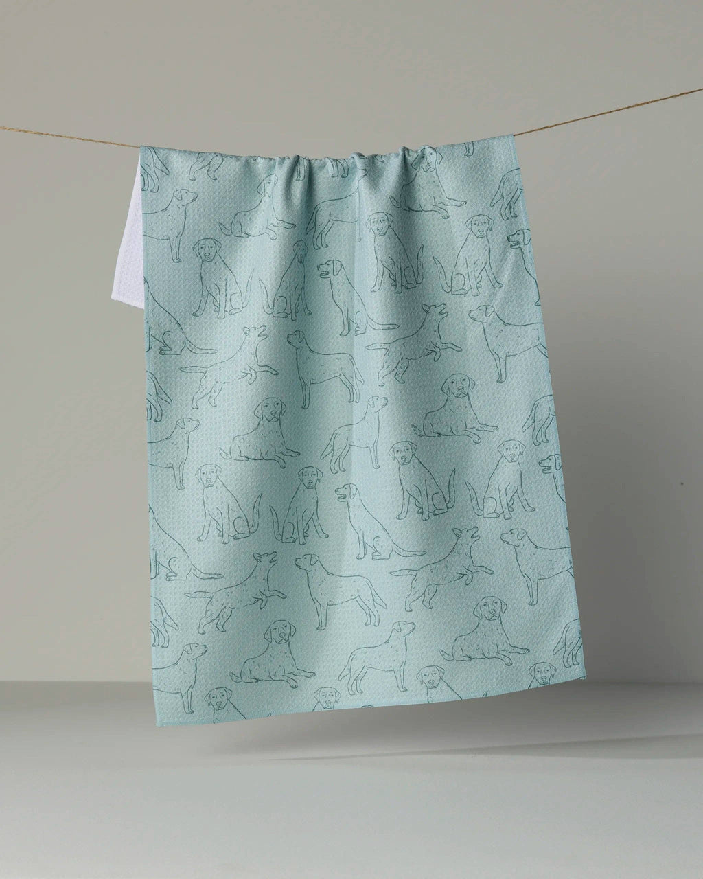 A light blue kitchen towel with a repeating pattern of dog line drawings, hanging from a string against a neutral gray background.