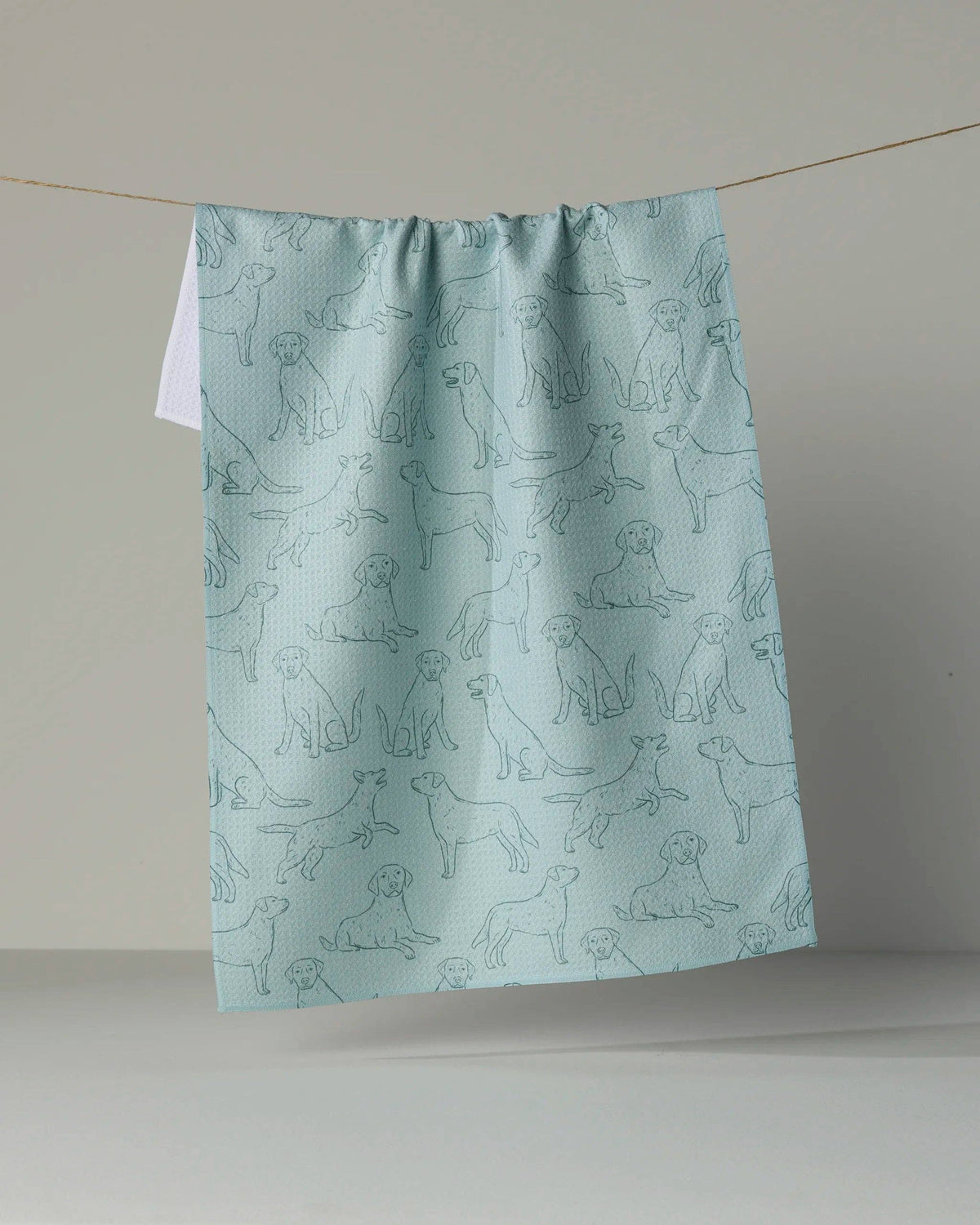 A light blue kitchen towel with a repeating pattern of dog line drawings, hanging from a string against a neutral gray background.