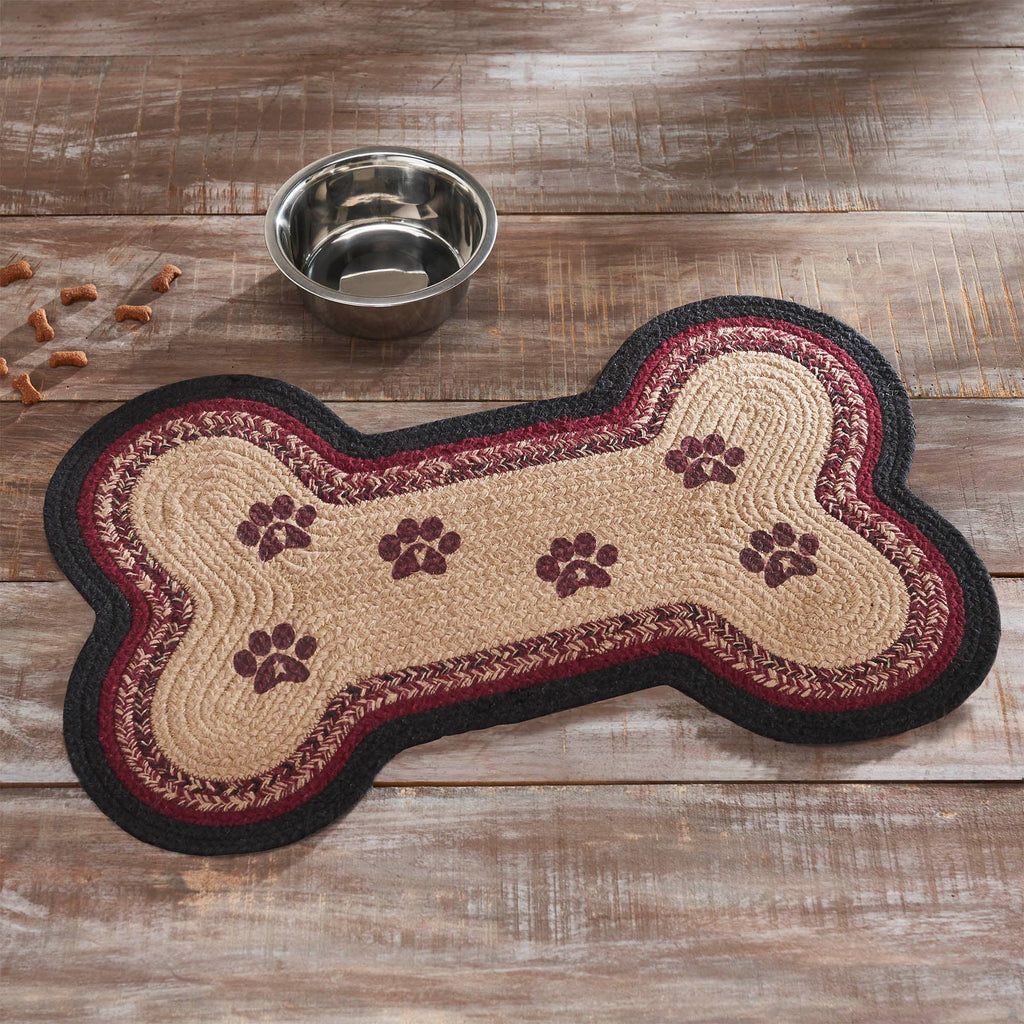 A bone-shaped braided rug with a beige center, dark red patterned border, and black outer edge, displayed on a rustic wooden surface next to a stainless steel bowl.
