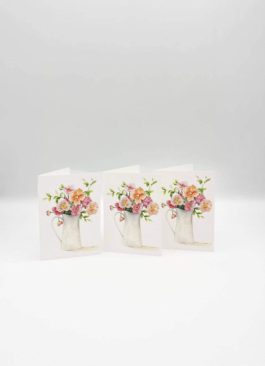 Three white greeting cards standing upright, each printed with a watercolor-style bouquet of pink, peach, and yellow flowers in a white pitcher, on a light gray background.