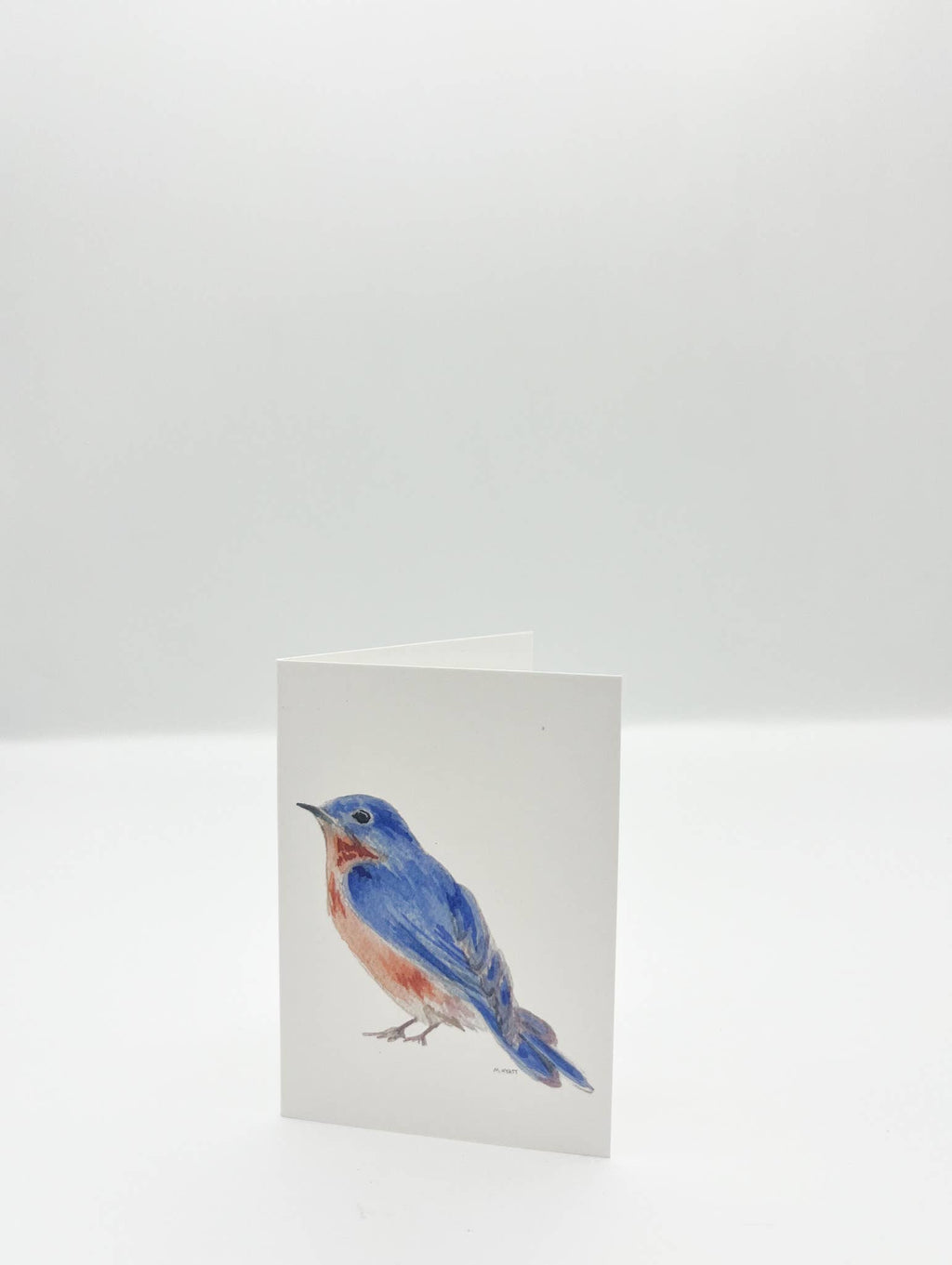 A folded white greeting card standing upright, printed with a watercolor-style bluebird on the front, photographed against a clean white background.