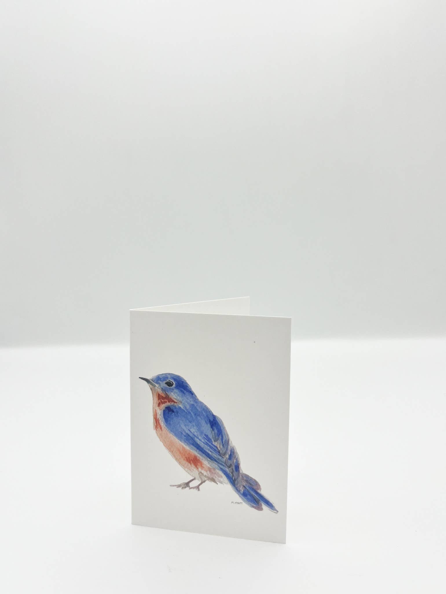 A folded white greeting card standing upright, printed with a watercolor-style bluebird on the front, photographed against a clean white background.
