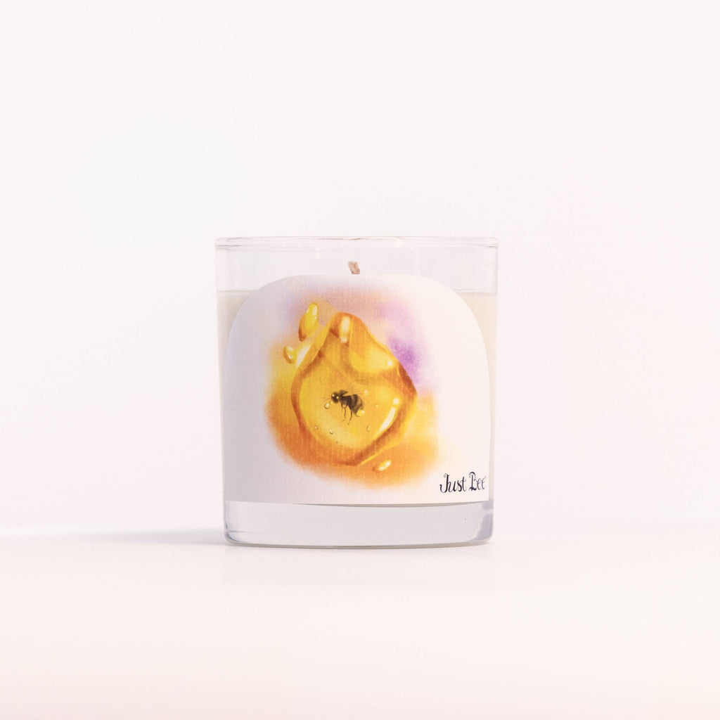 A clear glass jar candle with white wax and a printed label featuring a yellow-orange abstract flower or droplet design, photographed on a clean white background.