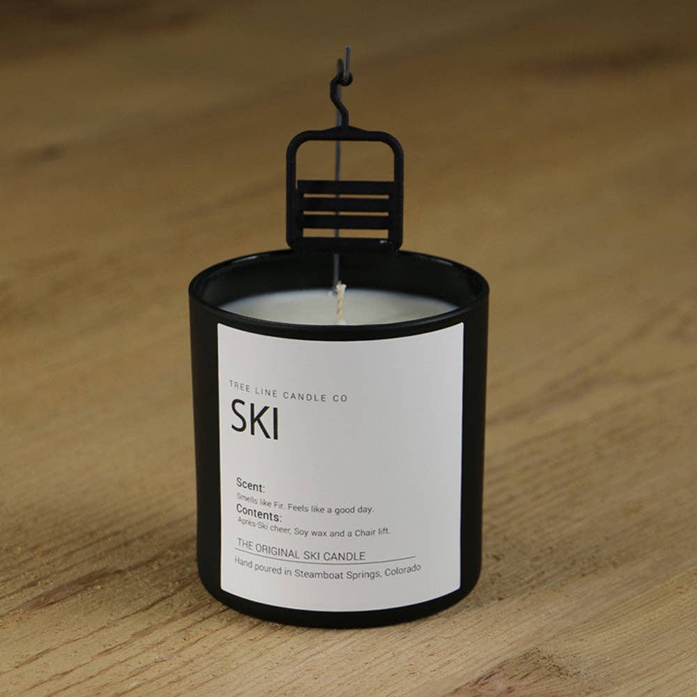 A black glass jar candle with a white label reading 'SKI' and 'TREE LINE CANDLE CO' on a light wood surface. A black metal wick holder sits on top of the candle.