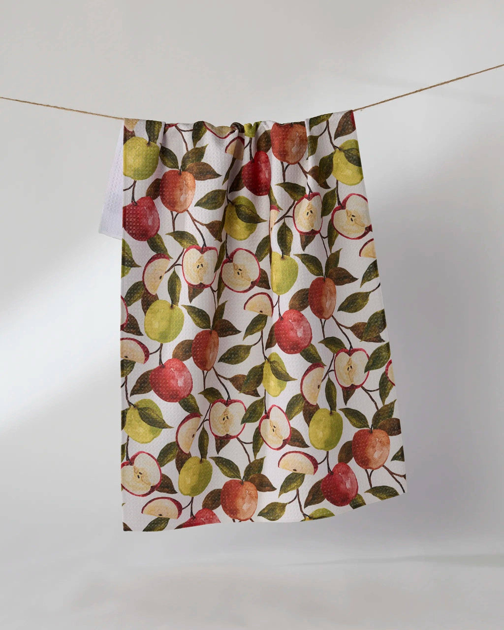 A kitchen towel with a watercolor-style apple pattern, hanging from a string against a plain light gray background.