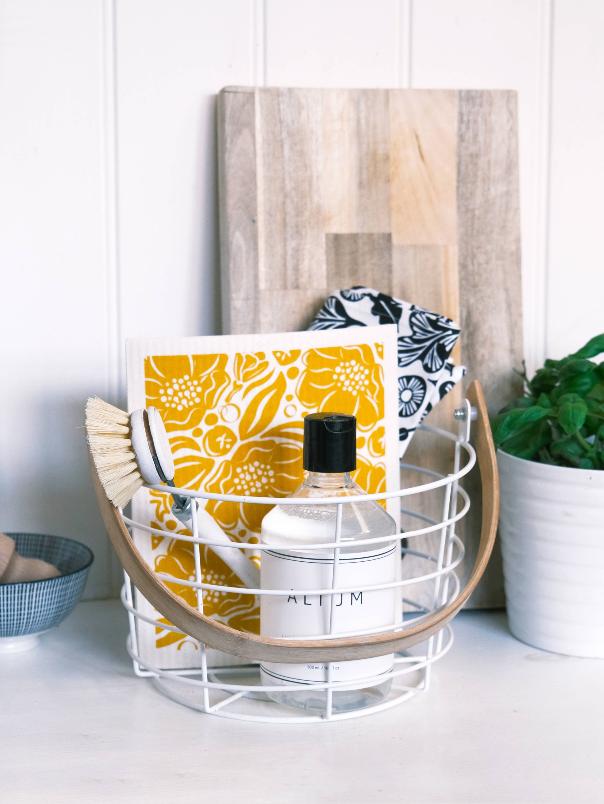 A white wire basket with a light wood handle sits on a countertop, styled with a yellow floral card, a white bottle, a scrubber, and a black-and-white patterned cloth. A wooden cutting board leans behind it against a white paneled wall, with a plant and bowl at the sides.