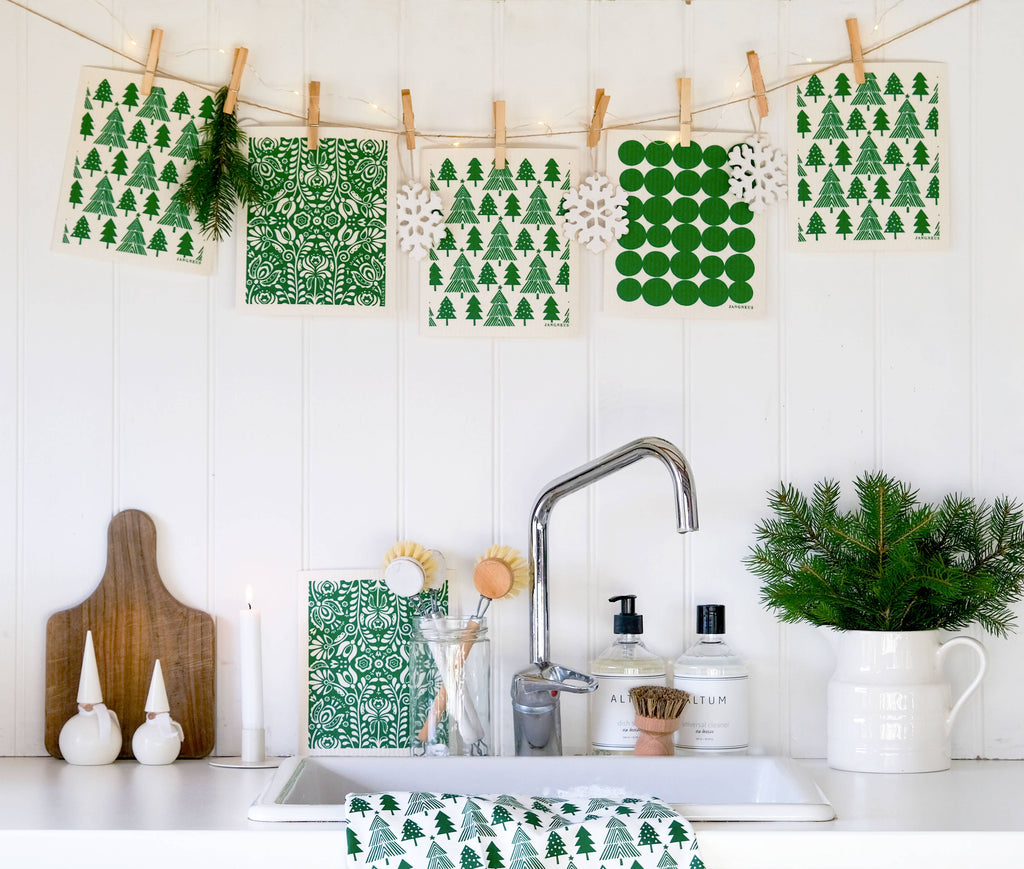 A styled kitchen scene with a white sink, faucet, soap dispensers, and a green-and-white patterned towel draped over the sink. Above the sink, fabric napkins or paper banners with green tree and snowflake patterns hang from a string with wooden clips.