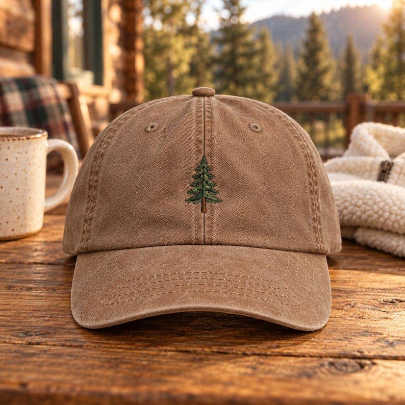 A brown washed cotton baseball cap with an embroidered green pine tree on the front, resting on a rustic wooden table with a mug and cozy blanket nearby, set against a blurred mountain forest background.