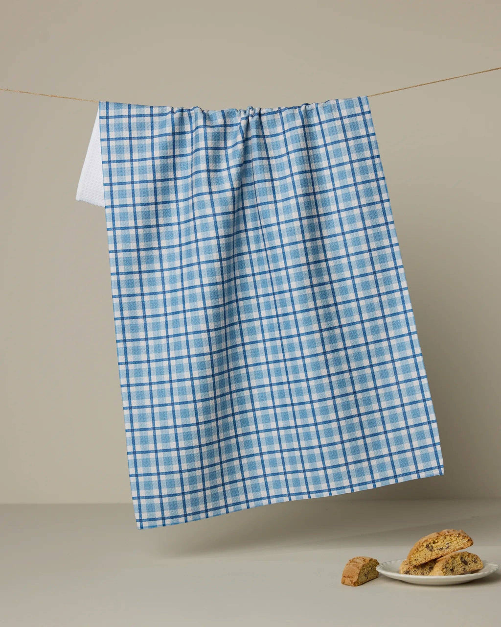 A blue and white plaid kitchen towel hangs from a string on a beige wall, with a small plate of cookies on a surface below.