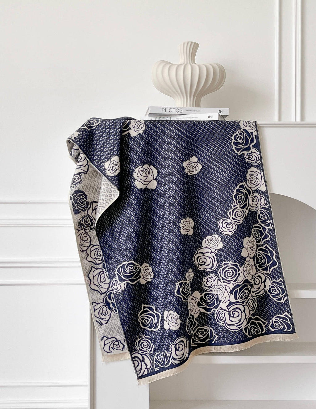 A navy blue throw blanket with a cream rose pattern and a textured geometric grid design, draped over a white mantel next to a decorative ceramic vase.