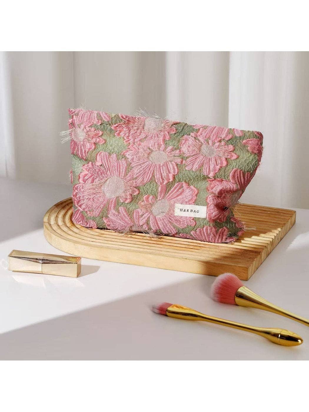 A pink and green floral embroidered makeup pouch with a small white 'MAYRAE' label, displayed on a wooden stand with gold makeup brushes and a gold compact on a white surface.
