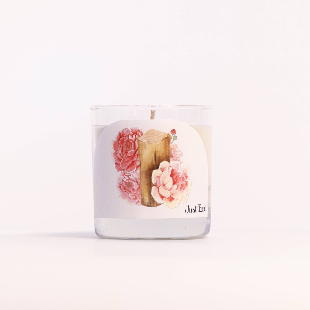 A clear glass jar candle with white wax and a round white label featuring a watercolor illustration of pink flowers and a wooden slice, with the brand name 'Just E.