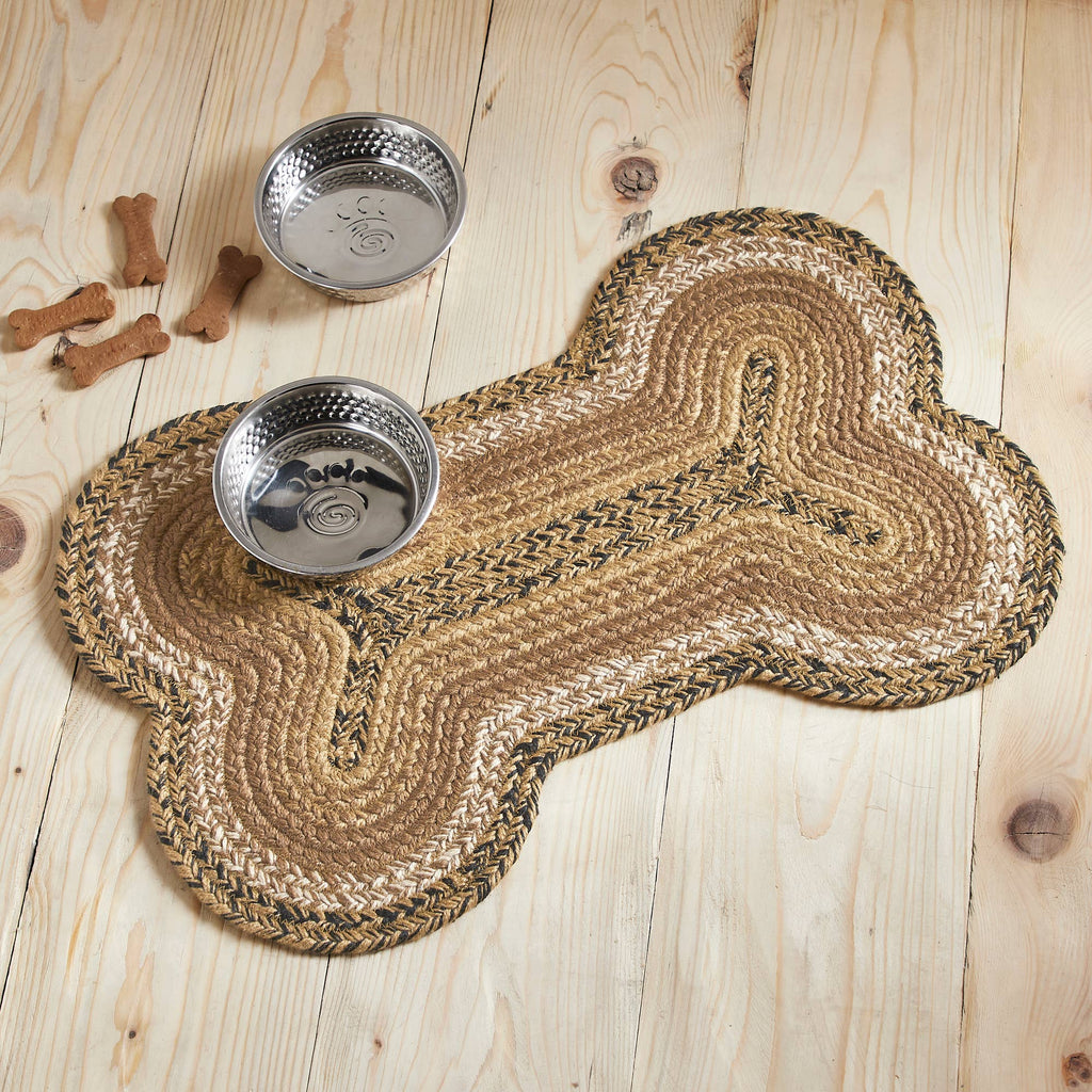 A braided bone-shaped pet mat in natural tan and brown tones, shown on a light wood floor with two metal bowls and a few dog bones as props.