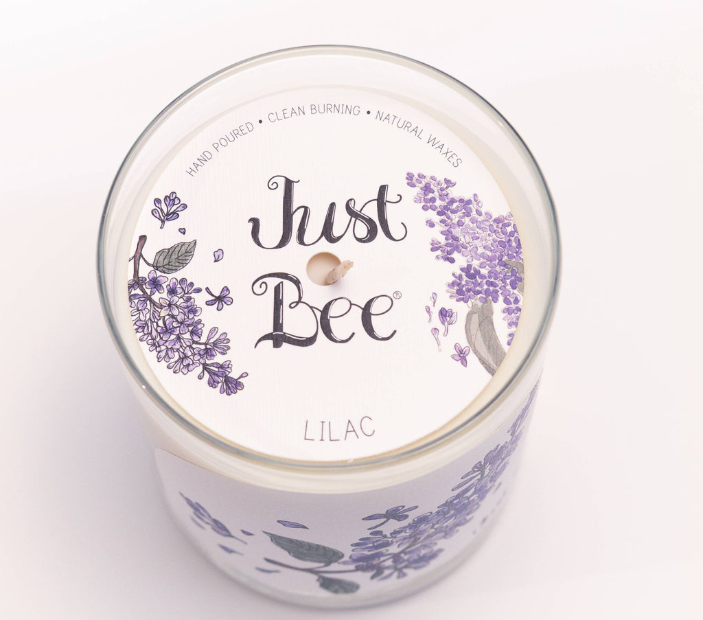A top-down view of a 'Just Bee' Lilac scented candle in a clear glass jar, featuring a white lid with purple lavender illustrations and the brand name 'Just Bee' in black script.