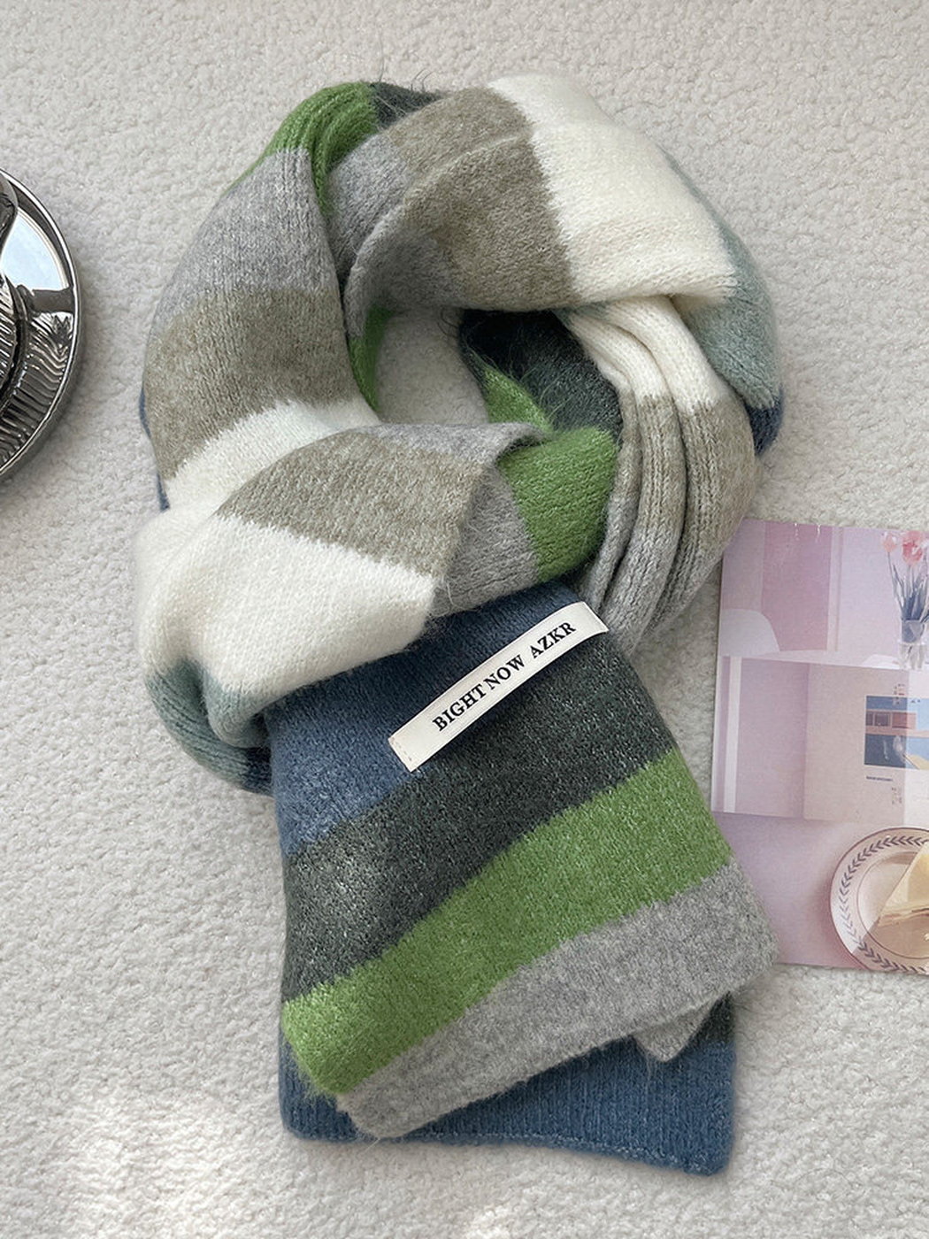 A chunky knit scarf with a geometric color-block pattern in green, gray, white, and blue, styled on a textured light gray surface next to a small card.