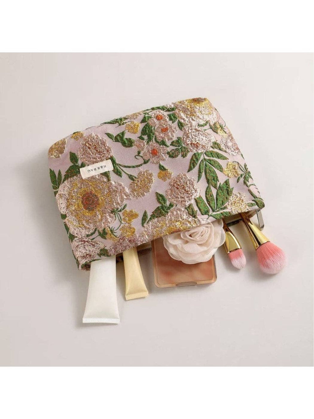 A floral makeup pouch with a pink base and embroidered yellow, white, and green flowers, shown with cosmetic items partially pulled out from the side on a light gray background.