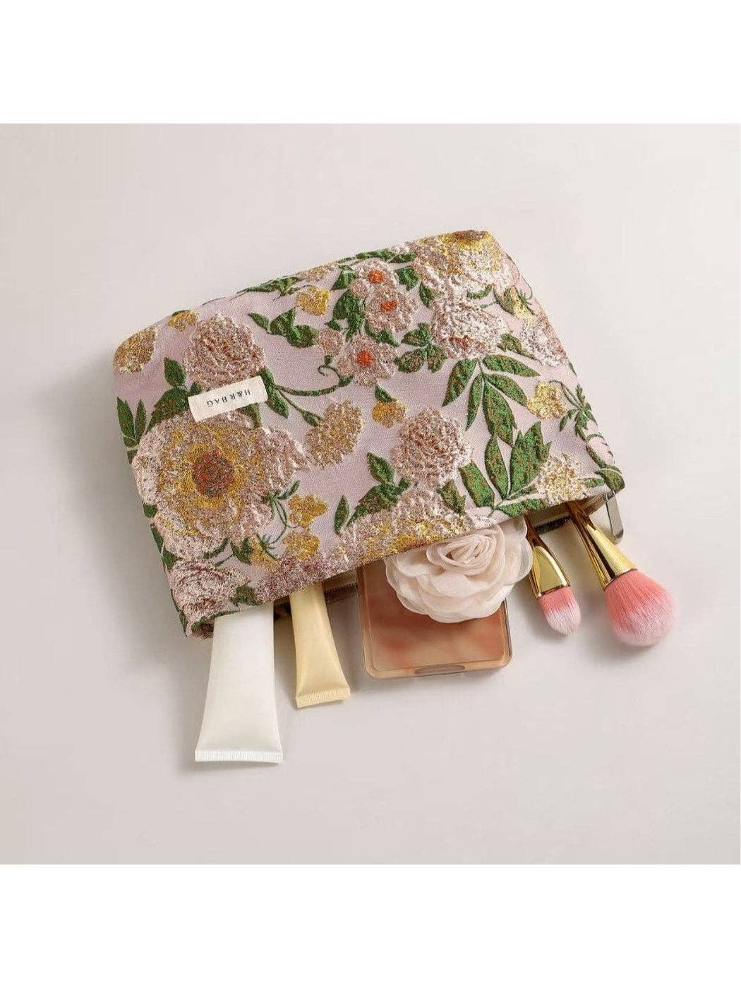 A floral makeup pouch with a pink base and embroidered yellow, white, and green flowers, shown with cosmetic items partially pulled out from the side on a light gray background.
