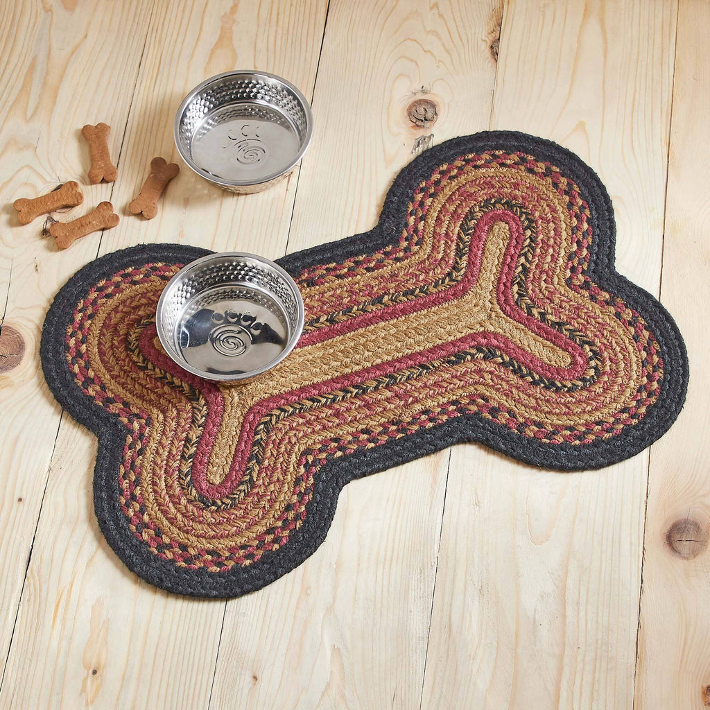 A braided bone-shaped pet mat in beige, brown, and burgundy with a dark gray border, shown on a light wood floor with two metal bowls and a few dog bones nearby.