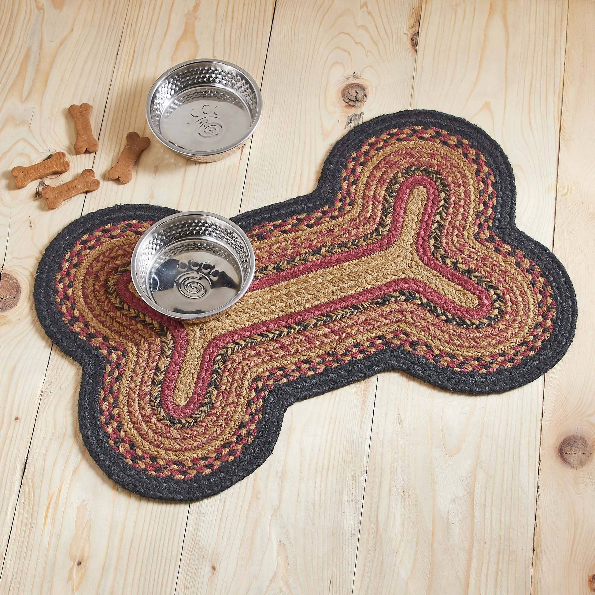 A braided bone-shaped pet mat in beige, brown, and burgundy with a dark gray border, shown on a light wood floor with two metal bowls and a few dog bones nearby.