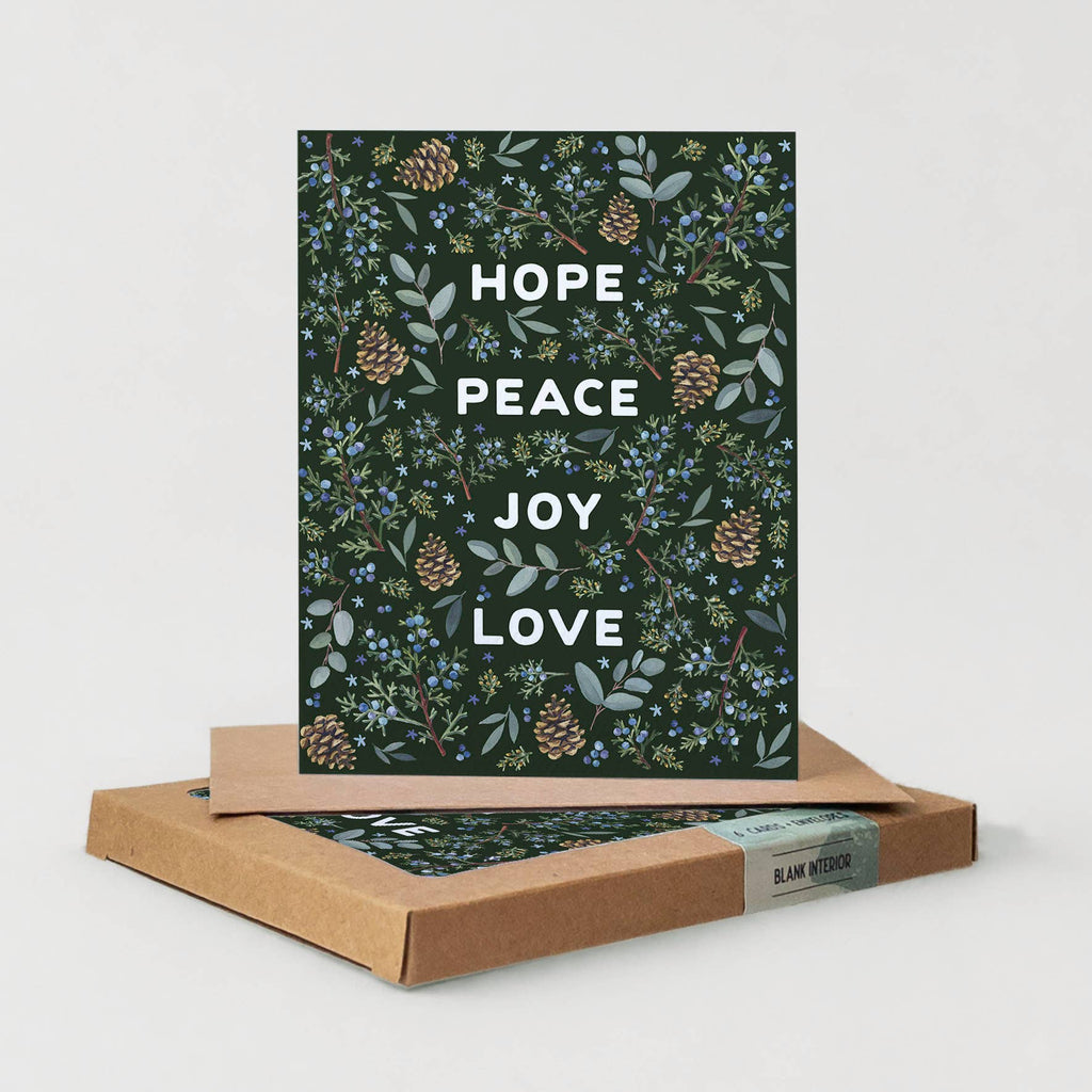 A greeting card with a dark forest green botanical pattern of pinecones, leaves, and blue berries, featuring the words 'HOPE PEACE JOY LOVE' in white. The card is displayed standing upright on top of its brown kraft paper packaging box against a light gray background.