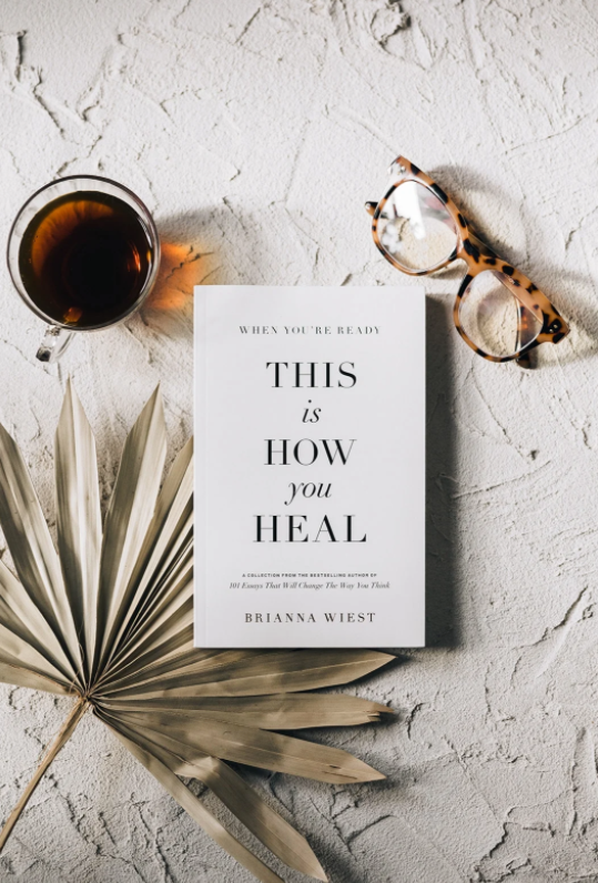 Book titled 'This is How You Heal' by Brianna Wiest on a textured surface with a cup of tea and glasses.