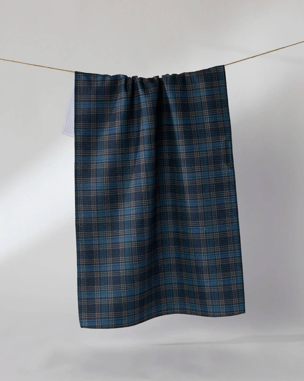 A dark blue and black plaid kitchen towel hanging from a thin wire against a light gray background.