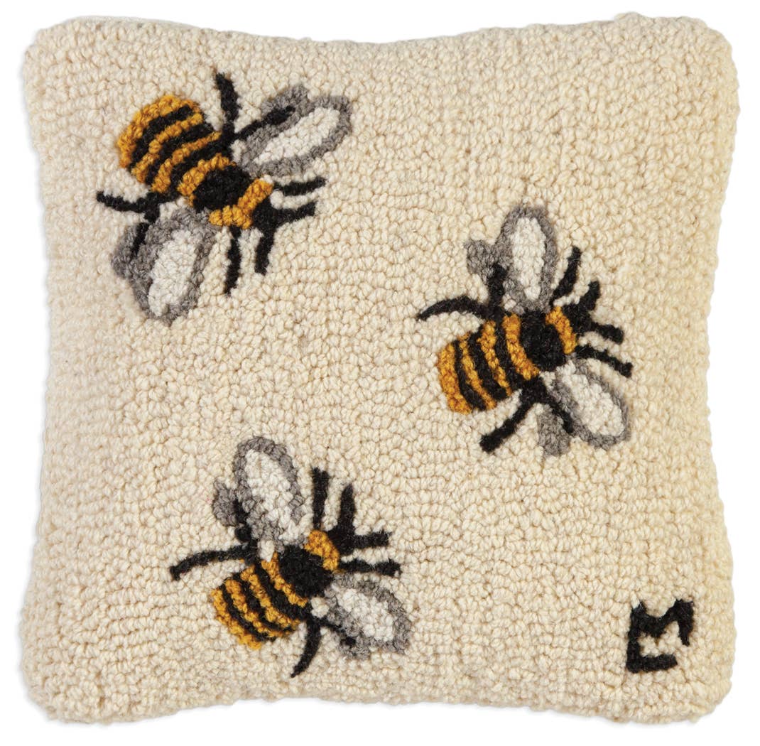 A square beige textured rug featuring three embroidered bumblebees in yellow, black, and white, with a small black monogram in the bottom right corner.