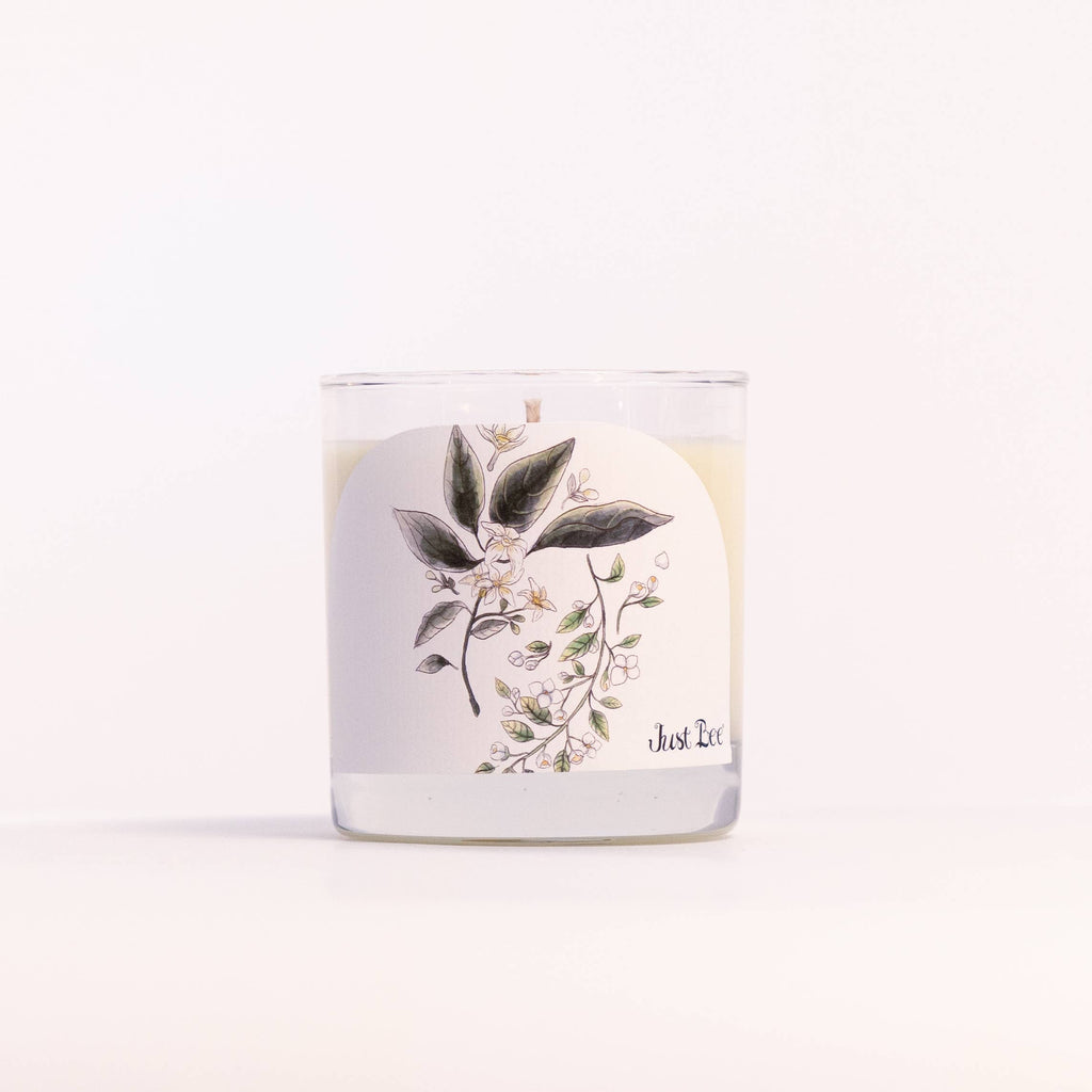 A clear glass jar candle with white wax and a white label featuring a botanical illustration of green leaves and small white flowers. The label has partial text reading 'Just Be' near the bottom right, set against a clean white background.