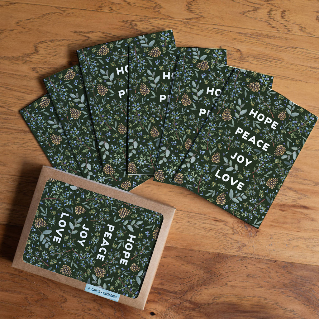 A set of square greeting cards with a dark green floral and pinecone pattern, fanned out on a wooden tabletop, with one card in a kraft envelope at the bottom left. The cards feature the words HOPE, PEACE, JOY, and LOVE in white.