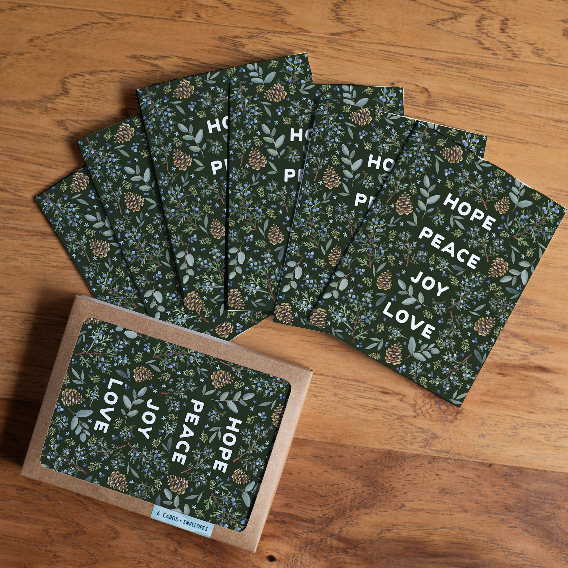 A set of square greeting cards with a dark green floral and pinecone pattern, fanned out on a wooden tabletop, with one card in a kraft envelope at the bottom left. The cards feature the words HOPE, PEACE, JOY, and LOVE in white.