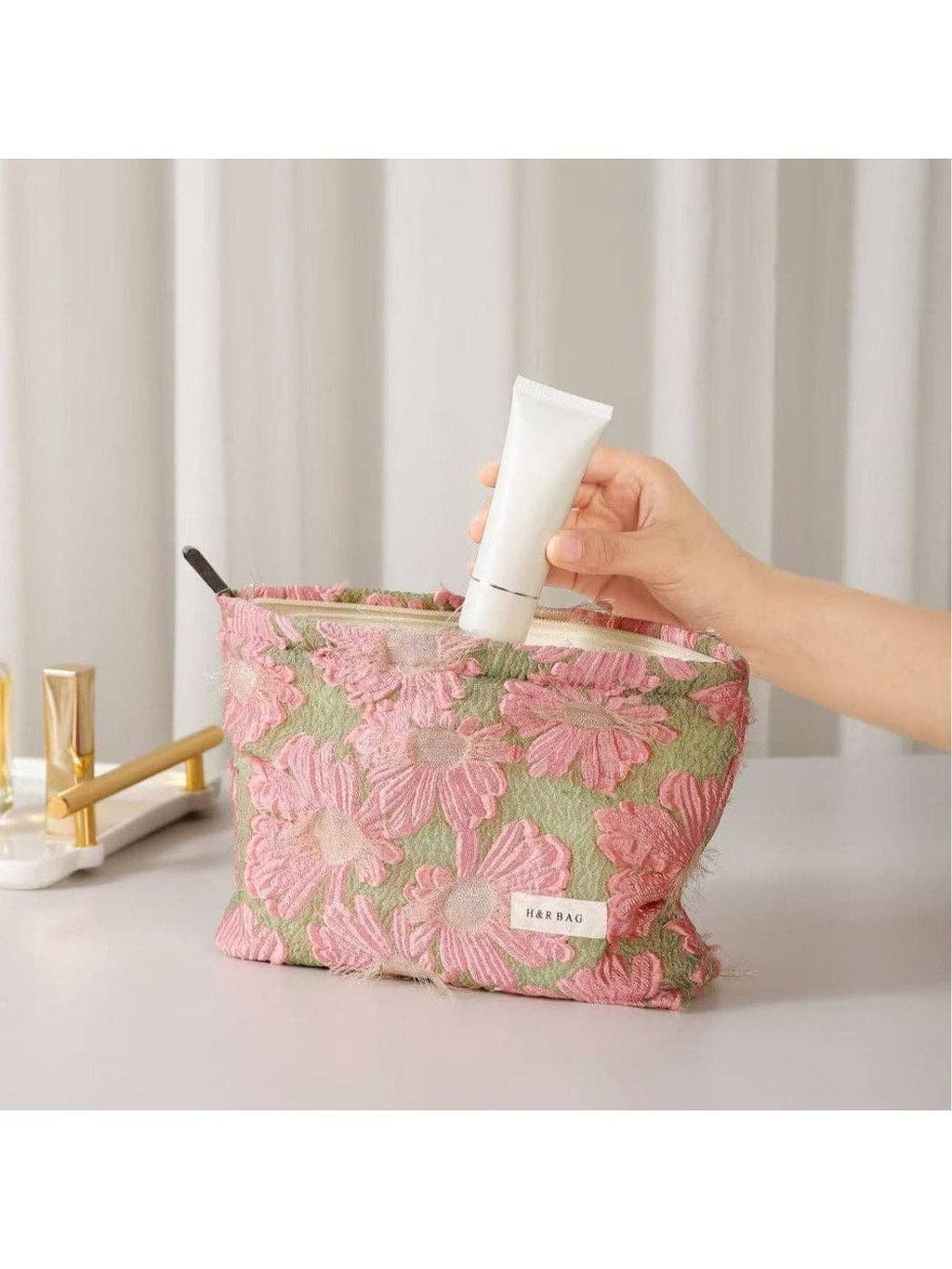 Hand holding a white tube of cream above a floral-patterned bag on a light surface with a neutral background