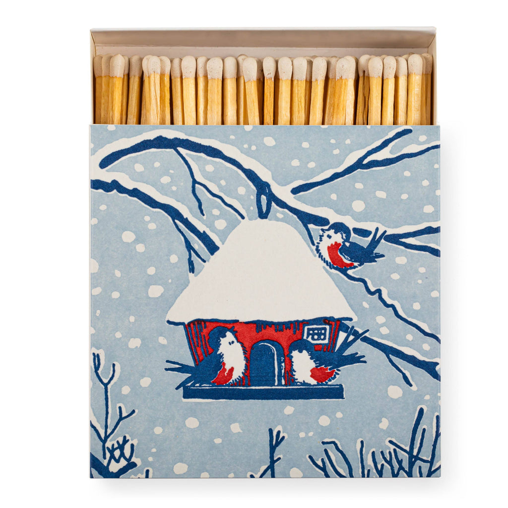 A matchbox with a winter birdhouse illustration on the front, shown open with wooden matches visible. The box features a light blue snowy background with dark blue branches, white birdhouse details, and red accents, plus small birds.