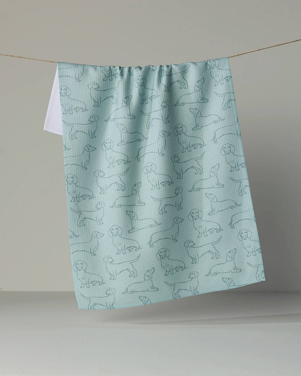 A light blue kitchen towel with a repeating pattern of dachshund dog silhouettes, hanging from a string against a neutral gray background.