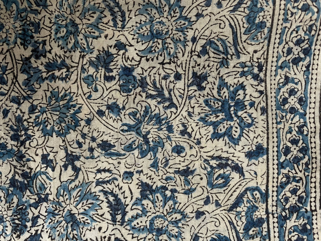 A close-up of a patterned textile featuring a blue and black floral and vine motif on a warm cream background, with a decorative border visible along the right edge.