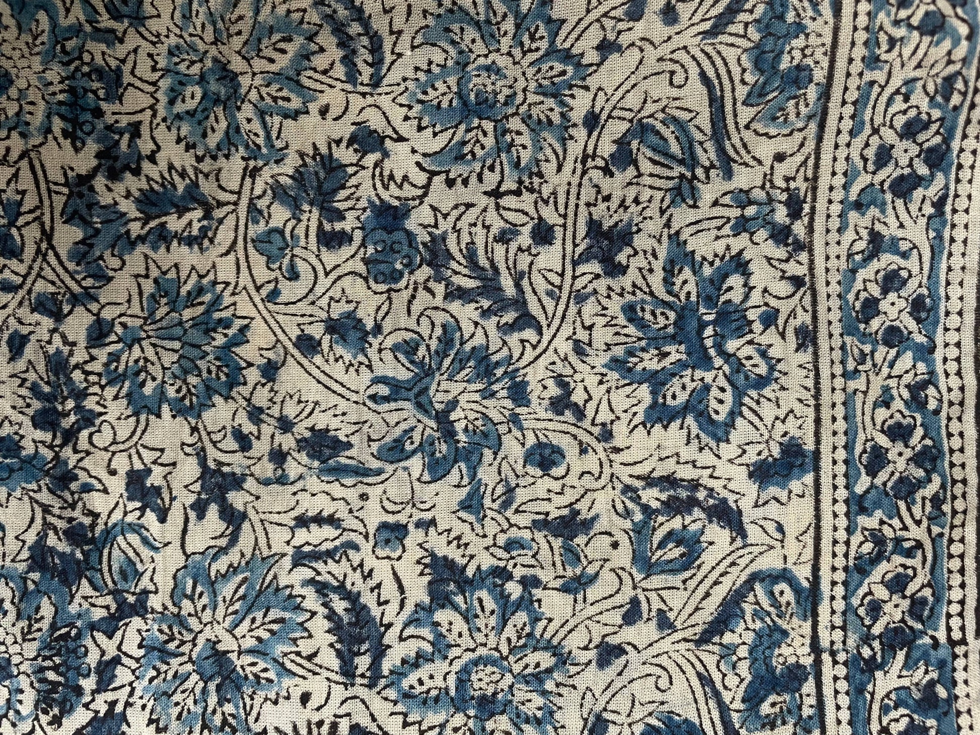 A close-up of a patterned textile featuring a blue and black floral and vine motif on a warm cream background, with a decorative border visible along the right edge.