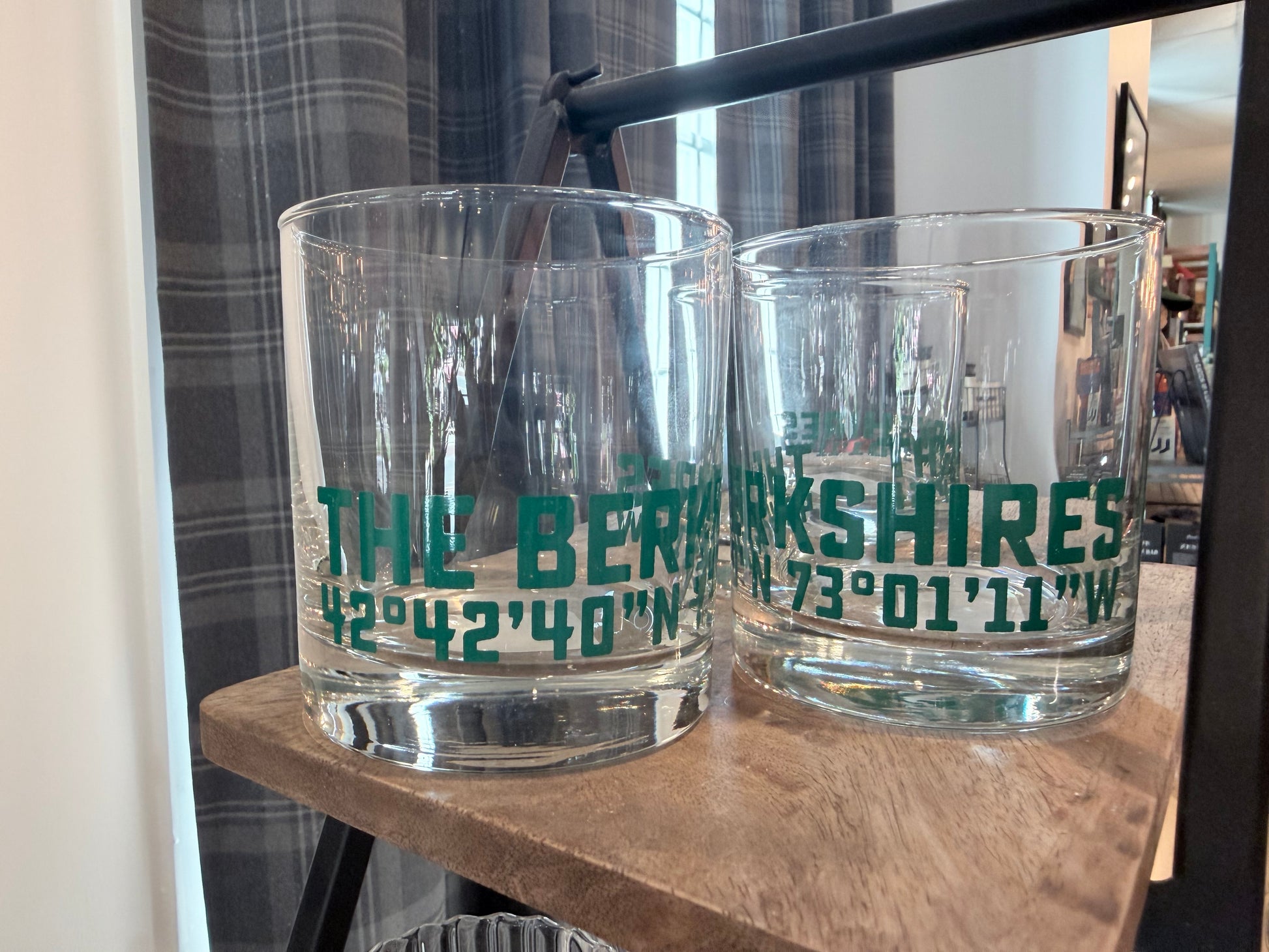 Two clear glass tumblers with green printed text reading 'THE BERKSHIRES' and coordinates, displayed on a wooden shelf indoors.