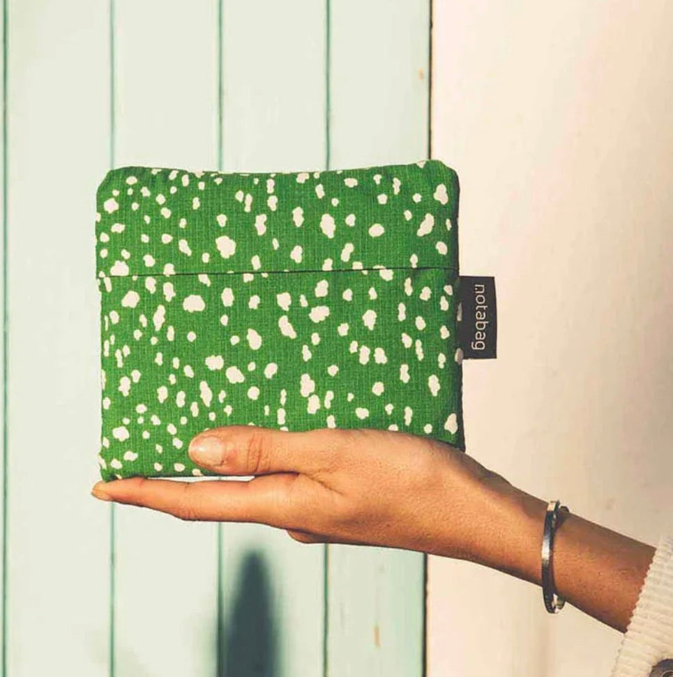 A hand holding a green fabric pouch with a white speckled pattern, featuring a black 'noobbag' brand tag, set against a background of light green vertical wooden planks and a cream wall.
