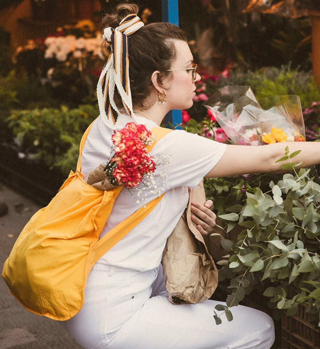 A woman in a white t-shirt and white pants is seen from behind, wearing a bright yellow drawstring backpack and carrying a brown paper bag. She is holding a small bouquet of red flowers and reaching toward flowers in a garden setting.