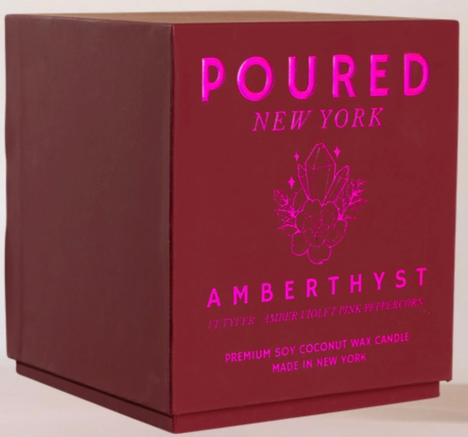 A square dark red or burgundy product box with bright pink text reading 'POURED NEW YORK' and 'AMBERTHYST,' plus a crystal-and-floral line drawing, photographed on a plain light background.