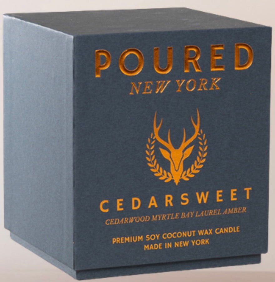 A dark slate blue square box for a Poured New York Cedarsweet soy coconut wax candle, featuring gold foil lettering and a deer head logo.