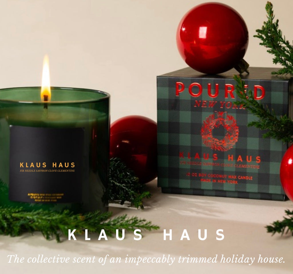 A Klaus Haus candle in a dark green glass jar with a lit flame, shown beside its branded box. The box is dark green plaid with red lettering and sits on a tabletop with red ornaments and evergreen branches around it.