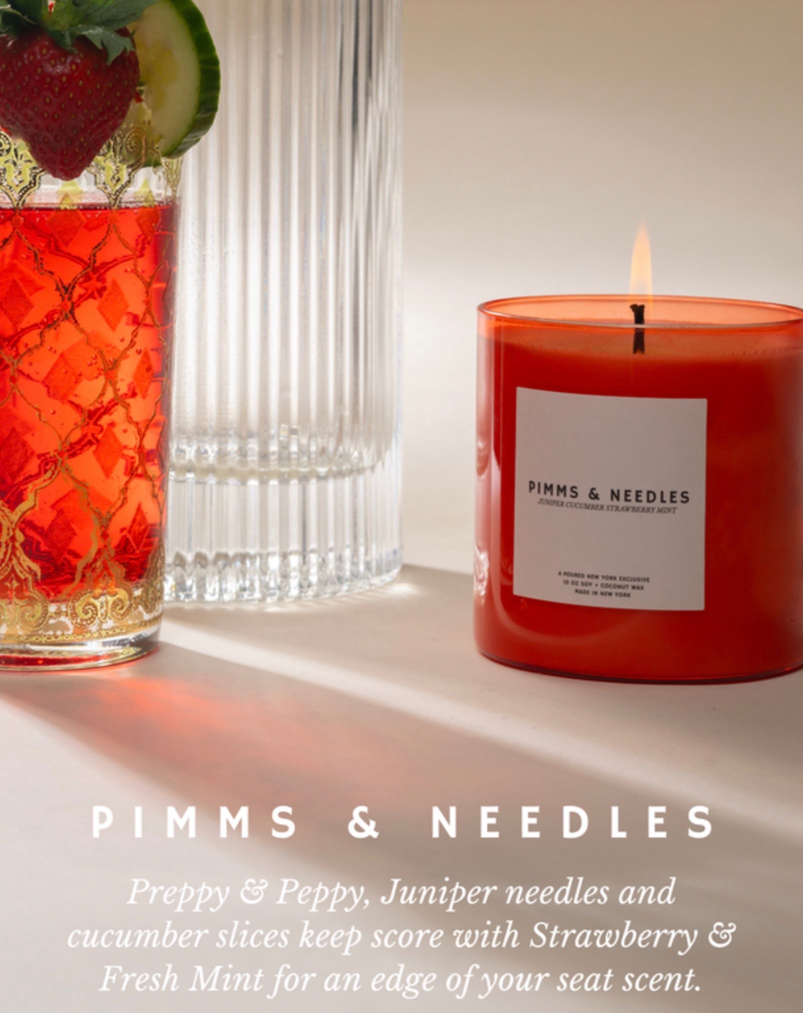 A red pillar candle labeled 'PIMMS & NEEDLES' sits on a tabletop beside a glass of pink-red cocktail garnished with a strawberry and cucumber, with a second ribbed glass behind it. Text at the bottom reads 'PIMMS & NEEDLES' and describes the scent as Preppy & Peppy, Juniper needles and cucumber slices keep score with Strawberry & Fresh Mint for an edge of your seat scent.