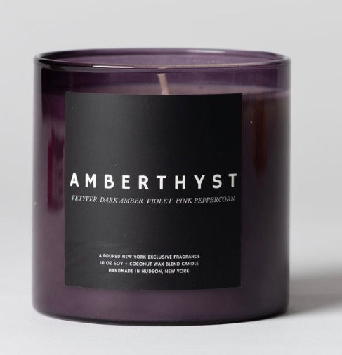 A dark purple glass jar candle with a black label reading 'AMBERTHYST' in white text, photographed against a light gray background.