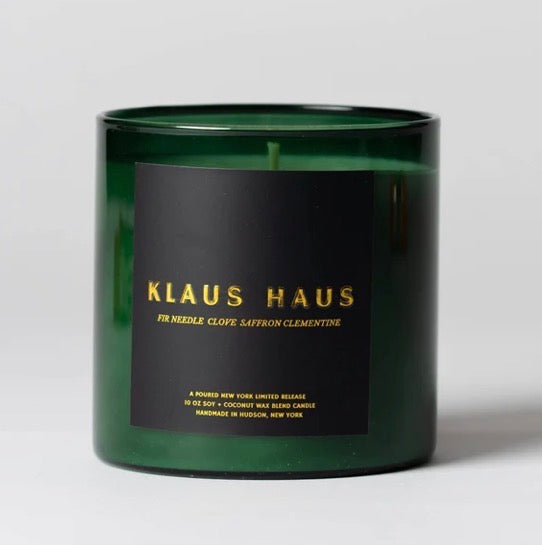 A dark green glass jar candle with a black label featuring gold lettering that reads 'KLAUS HAUS'.