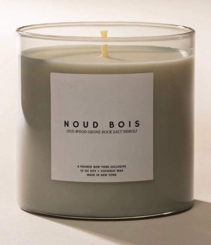 A large glass jar candle with a single wick, filled with light warm gray wax. The front features a large white square label with the text 'NOUD BOIS' and smaller scent details.