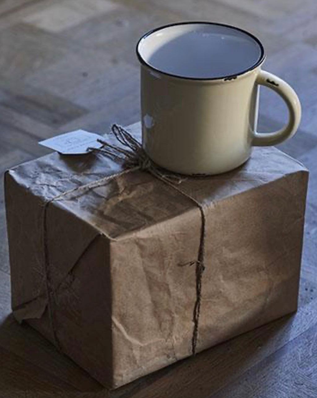 An off-white enamel mug with a black rim sits on top of a brown paper-wrapped gift box tied with twine on a wooden floor.