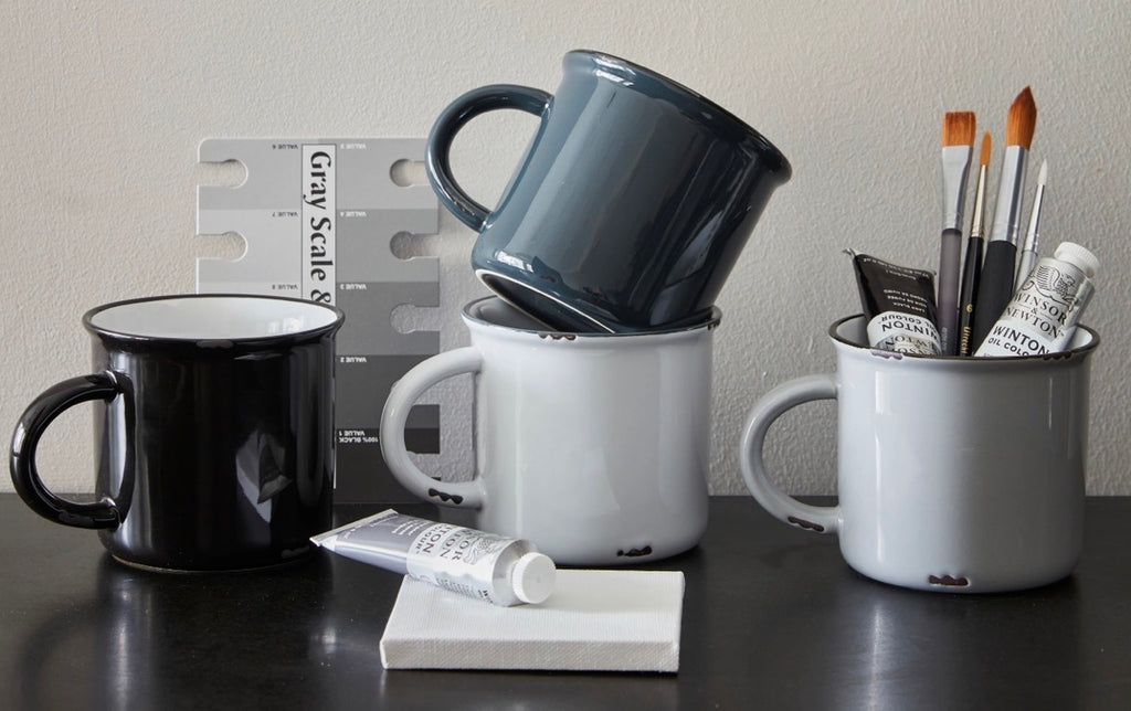 A collection of ceramic mugs and paint storage cups arranged on a dark tabletop against a light wall. The set includes a black mug, a dark blue-gray mug stacked on top of a white enamel mug, a white enamel mug holding paintbrushes, and a tube of white paint on a small white block.