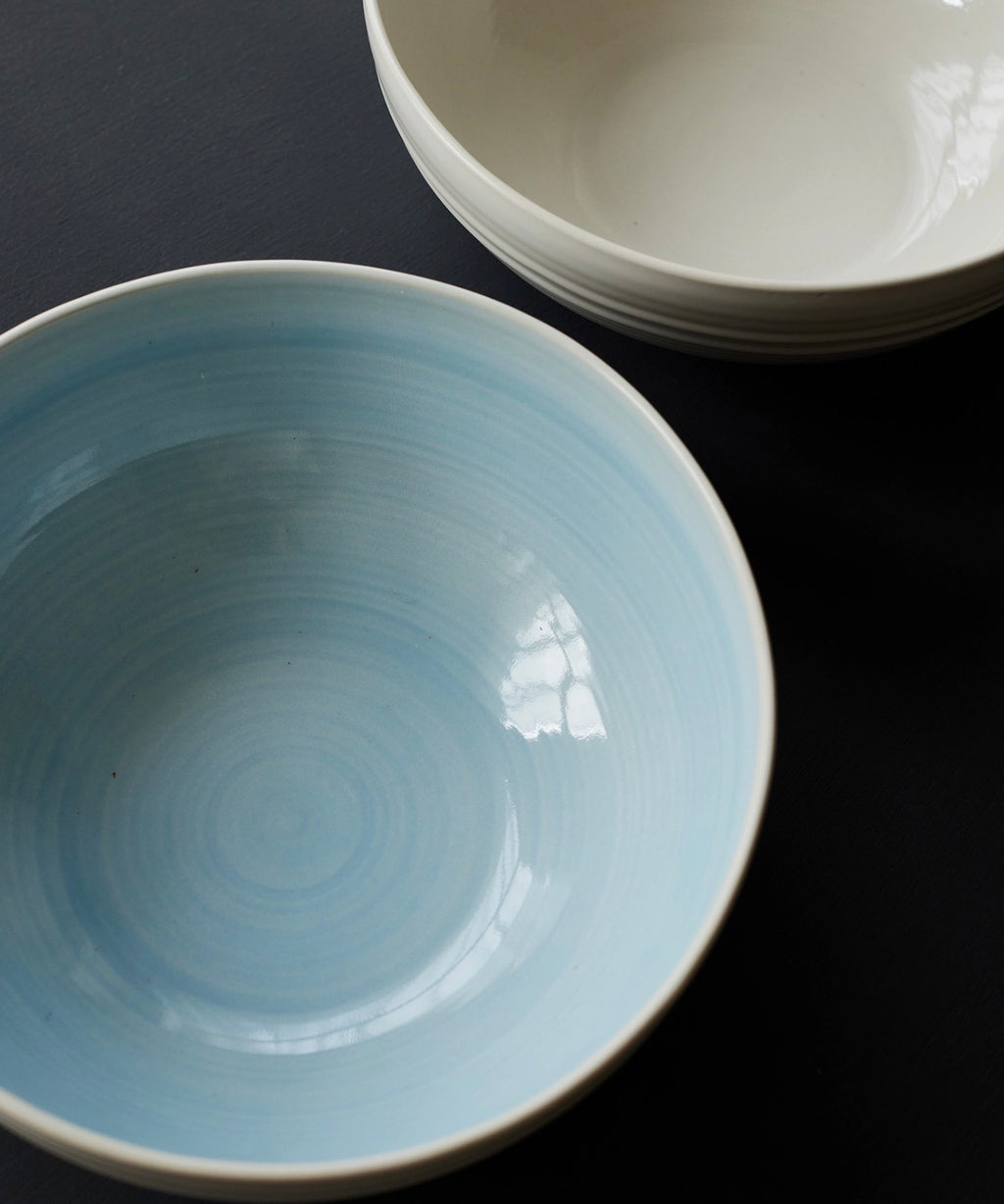 A light blue glazed ceramic bowl in the foreground on a dark background, with a stack of white bowls partially visible in the upper right.